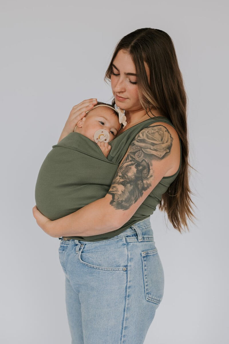 Frau mit Baby in grünem Tragetuch, Tattoo am Arm, lässige Jeans, stillt liebevoll.