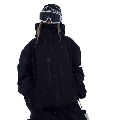 Person in schwarzer Schneejacke mit Stehkragen, Mütze und Skibrille, Frontansicht im Schneefall mit locker fallendem, weitem Schnitt. 3