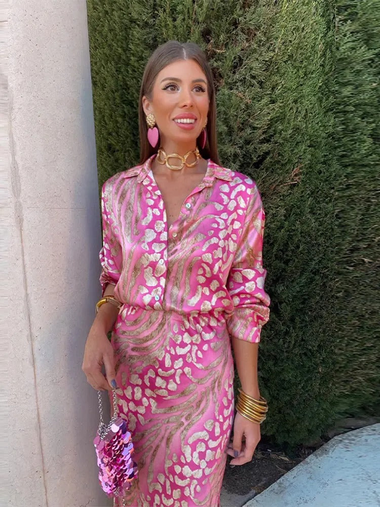 Frau in pinkem, gemustertem Kleid mit goldenen Accessoires vor grüner Hecke.
