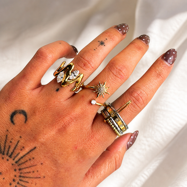 Hand mit tätowierten Motiven trägt den goldfarbenen Wellenring mit zwei klaren Steinen zusammen mit weiteren Ringen auf weißem Stoff. 3