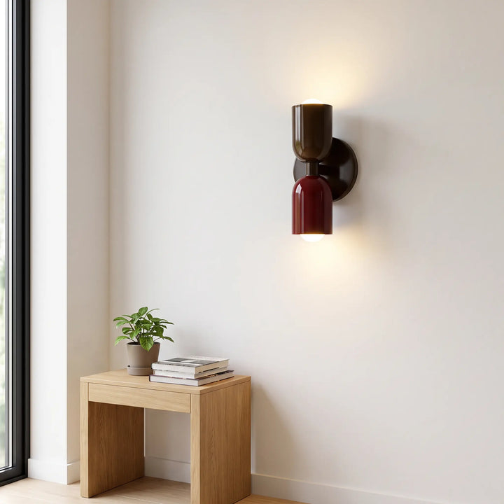 Wandleuchte mit dunkelbraunem oberem und weinrotem unterem Schirm an heller Wand über einem Holzhocker mit Pflanze und Büchern. 10