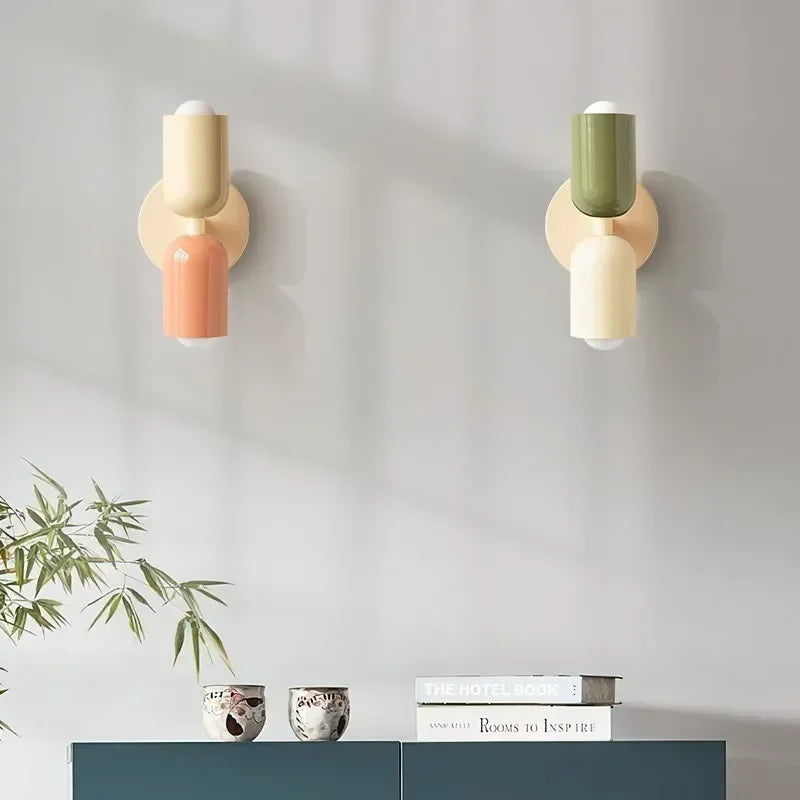 Zwei Wandleuchten nebeneinander an grauer Wand, links Creme mit Rosa, rechts Grün mit Creme, darunter ein Sideboard mit Büchern. 14