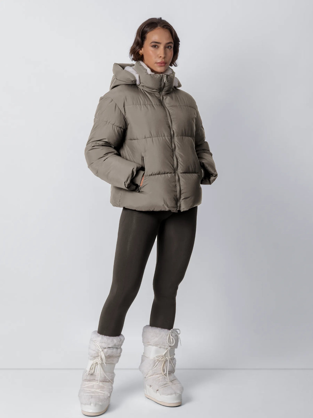 Ganzkörperansicht: Model trägt die graubraune Steppjacke mit Kapuze und weißem Fleecefutter, dazu dunkle Leggings und weiße 21