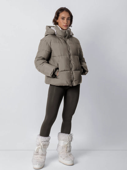 Ganzkörperansicht: Model trägt die graubraune Steppjacke mit Kapuze und weißem Fleecefutter, dazu dunkle Leggings und weiße 21