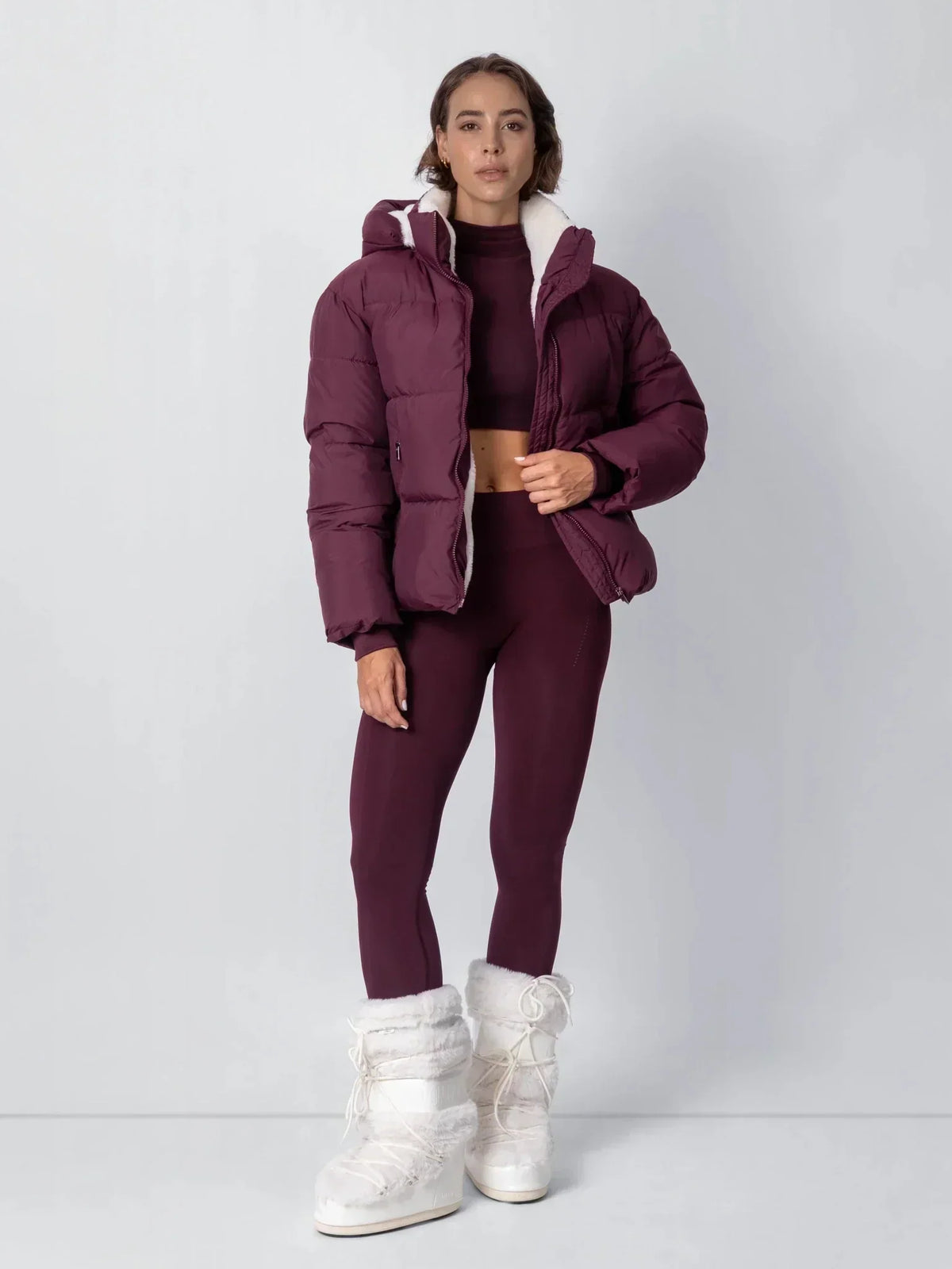 Ganzkörperansicht der beerenfarbenen Jacke mit Leggings und weißen Pelzstiefeln, Hand am Reißverschluss 17