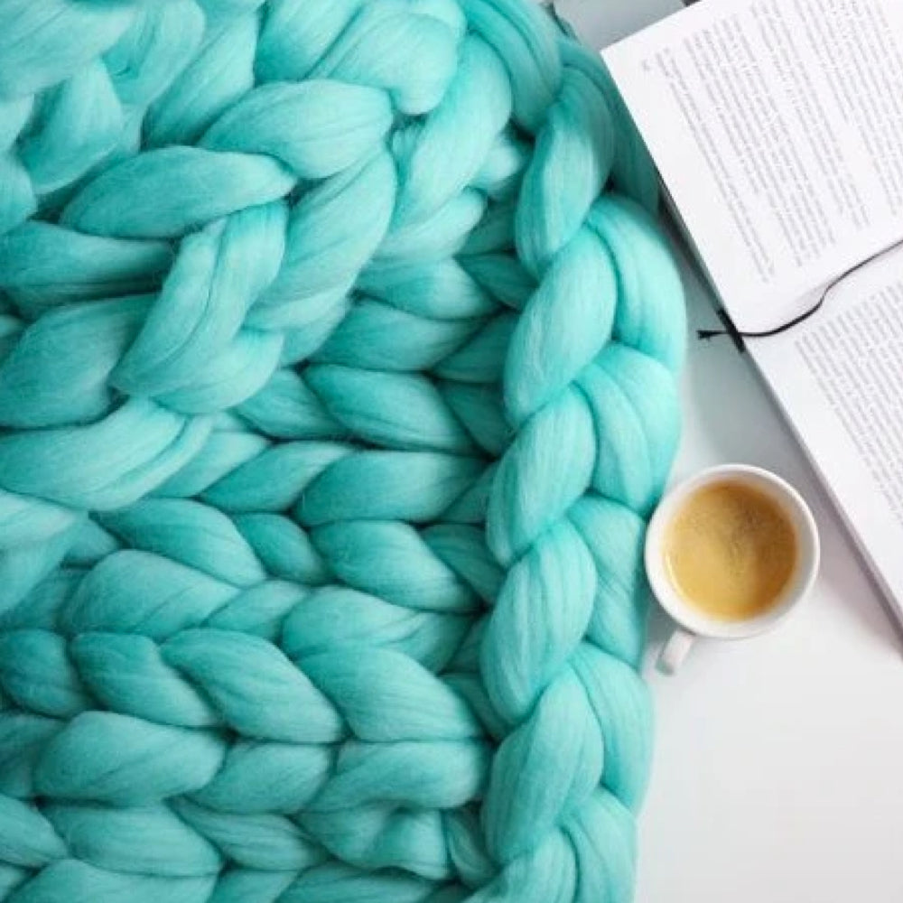 Türkise grob gestrickte Decke aus Wolle neben Buch und Kaffeetasse auf weißem Hintergrund.