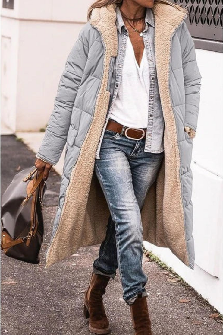 Frau trägt die hellgraue lange Steppjacke offen mit Teddyfutter, Jeansbluse, Gürtel und Stiefeln auf einem Gehweg, Handtasche in der Hand. 8