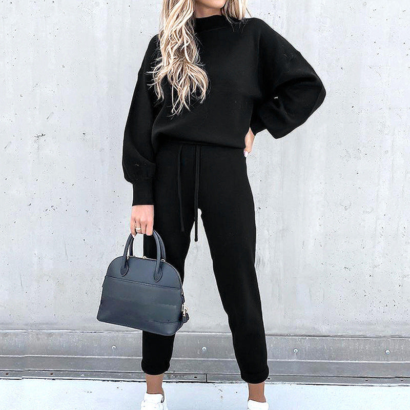 Frau in schwarzem Zweiteiler Hausanzug mit Rollkragen, High-Waist-Hose, hält schwarze Handtasche.