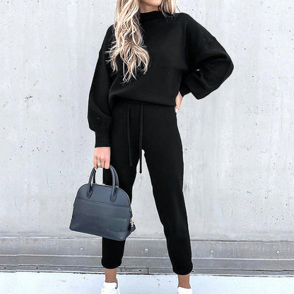 Frau in schwarzem Zweiteiler Hausanzug mit Rollkragen, High-Waist-Hose, hält schwarze Handtasche.