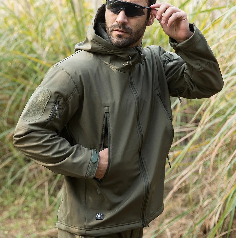 Model mit Sonnenbrille in der olivgrünen Jacke im hohen Gras, eine Hand in der Tasche, die andere an der Brille. 11